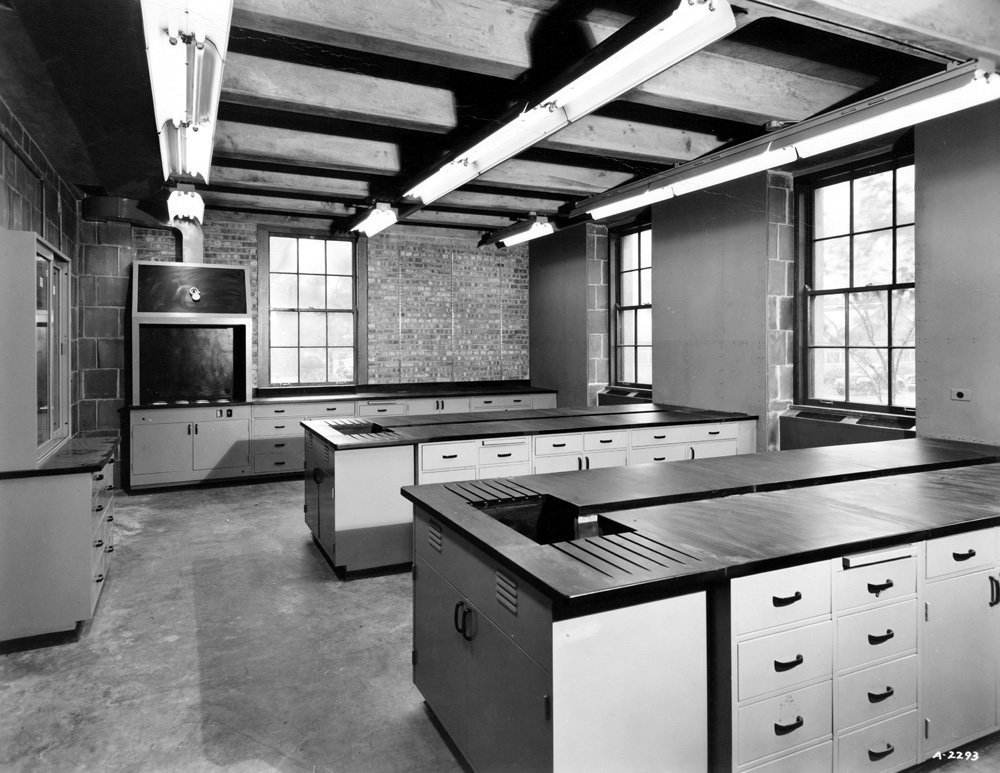 Black-and-white photograph of an empty laboratory in the American Meat Institute building on the University of Chicago campus. The room includes two lab benches in the center of the room, a bench and hood along one wall, and a cabinet along another wall. Cement floor, exposed brick walls, exposed beams with florescent lighting, and three windows.
