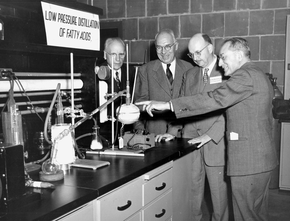 Black-and-white photograph of four middle-aged white men in suits looking at scientific equipment set up on a lab bench. A sign above the lab bench reads "Low pressure distillation of fatty acids."