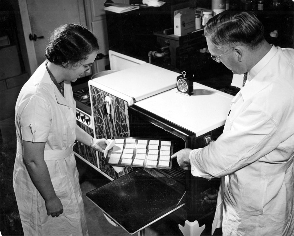 Black-and-white photograph of a middle-aged white man and a young white woman wearing white lab coats looking at a sheet pan of square pie crusts that appear to have just been removed from a small oven.