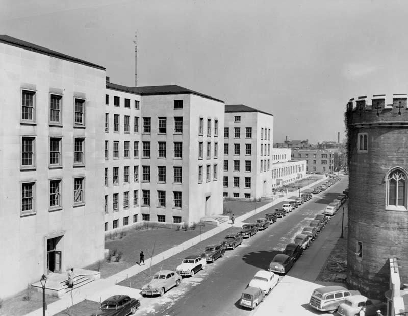 A black and white photo looking over a street lined with cars. On the left side of the street, there's a large, blocky building made of light colored stone with rows of square windows. On the right side of the street, part of a turreted tower is visible.