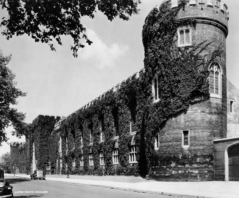 A black and white photo looking along an ivy covered wall and the turreted tower at the end of the wall.