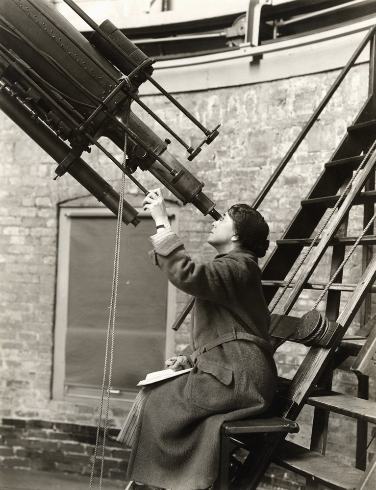 Black and white image of woman wearing a jacket with her dark hair pulled back. She is seated on a bench attached to a staircase, and is looking through the eyepiece of a large telescope. Her left hand is on one of the adjusting knobs of the telescope.