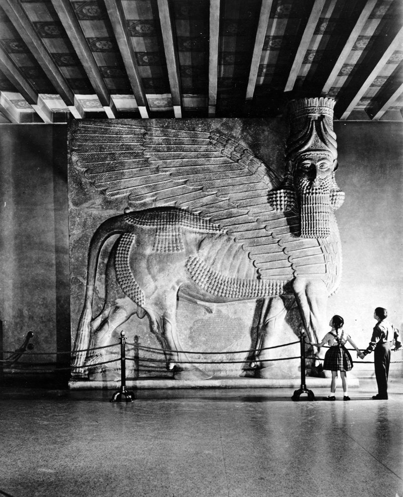 Photograph of the human-headed winged bull installed at the Oriental Institute. Two children look at the sculpture, and provide a sense of scale for the monumental work.