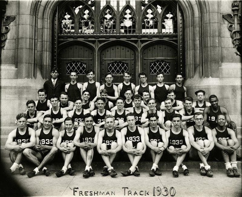 Forty men in track uniforms sit on the steps of a Gothic building, smiling for the camera.