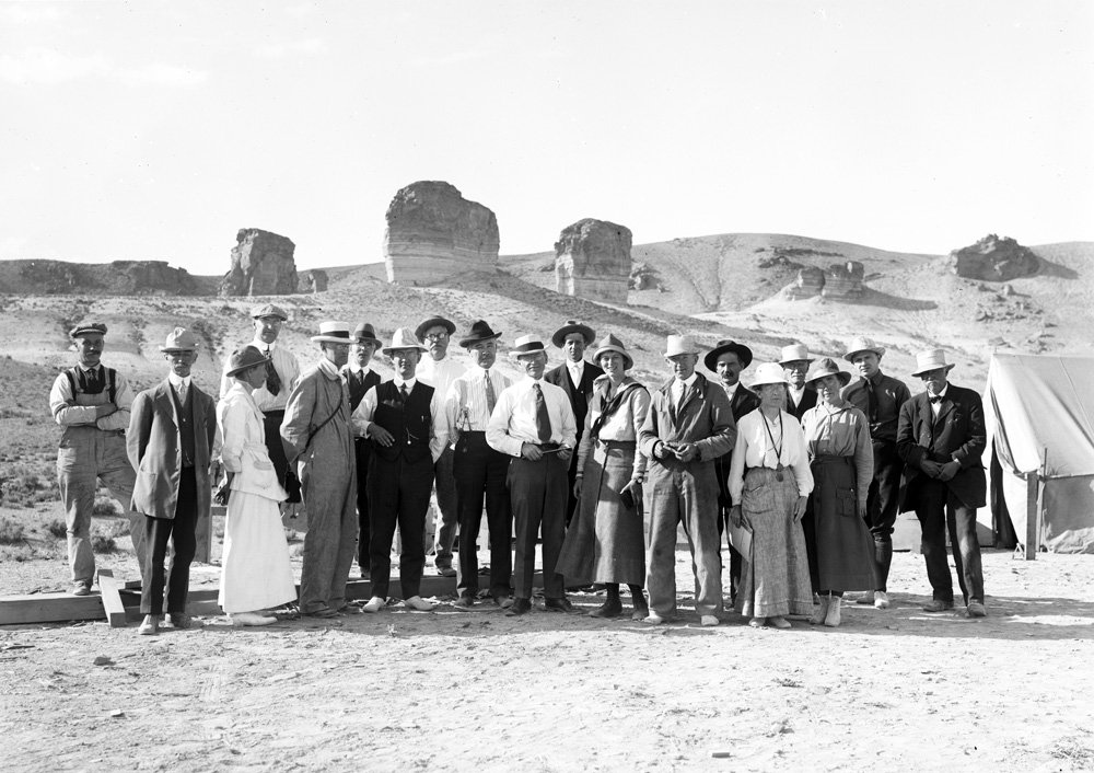 Black and white photo of large group of people standing in desert. Three large rock formation are behind them. All are wearing hats, and are dressed in a variety of clothing, including suits and ties, coveralls, and skirts and blouses.
