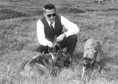 A black and white portrait of Dino Buzzati, squatting in a field with a dog.