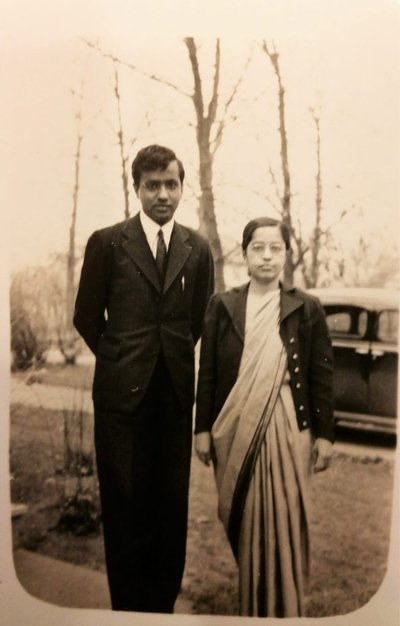 A suited man and a woman in robes, both wearing glasses, pose on a front lawn.