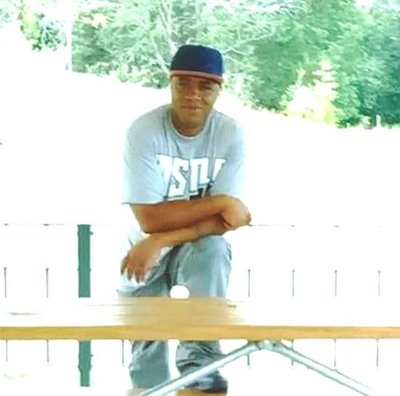 A middle aged person with medium skin tone, wearing a grey shirt and pants and a black baseball cap. He is standing behind a picnic table with one foot on the bench, resting his arms on his knee.