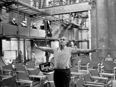 A black and white image of Luigi Nono standing with his arms outstretched in an empty room filled with chairs.