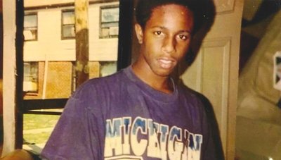 A Black man in his twenties, wearing a purple shirt, standing in front of a window. You can see an apartment building through the window.