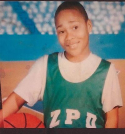 professional picture of Mr. Miller as a child. He is Black with medium skin tone and short hair and about 10 years old. He is in a white t-shirt and green basketball jersey that says ZPD. He is holding a basketball under his right arm.