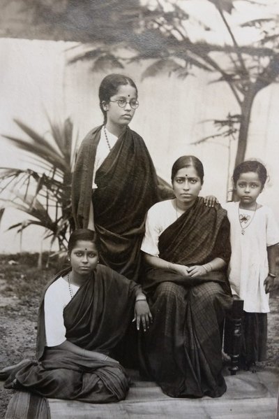 A woman in robes and glasses stands by two sitting women and one standing child.