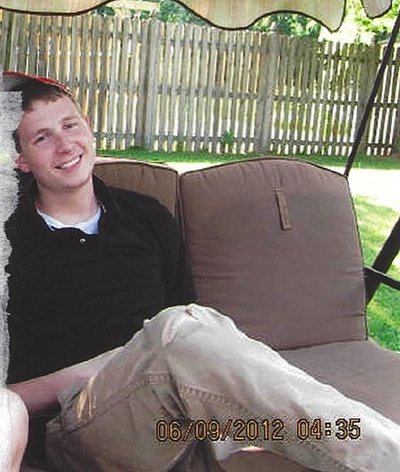 A white man in his twenties, wearing a black Polo shirt and khaki pants, sitting cross legged on a porch swing. There is a brown picket fence behind him.