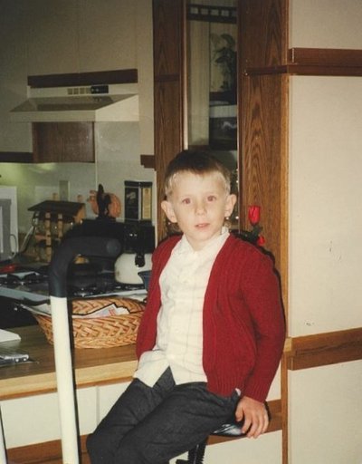 A white child, approximately 7 years old, sitting on a chair next to a kitchen counter, wearing a white shirt and red cardigan