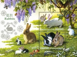 Brown, white, and black rabbits sit on green grass under a tree with purple flowers.