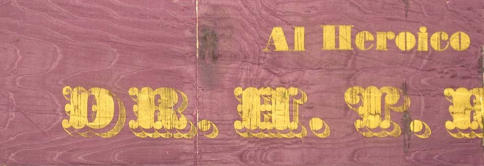 A dark purple sash with yellow woodcut letters printed on it. Text cut off, but shows "Al Heroico."
