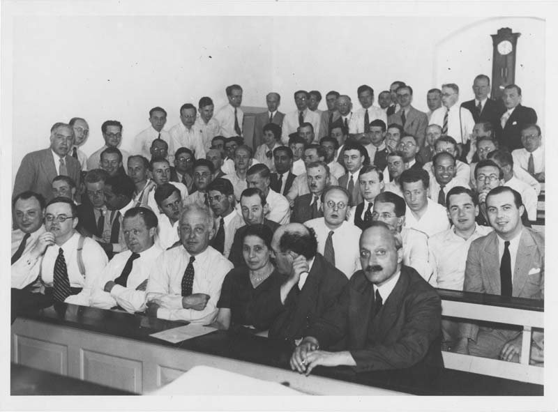 A black and white photo of men and one woman, sitting in the front row, in a lecture hall