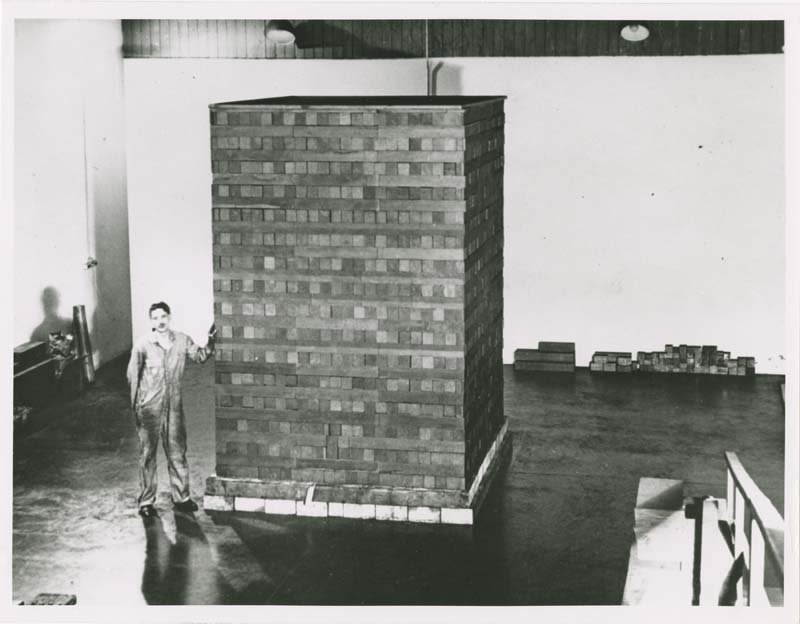 A black and white photo of a man standing next to the exponential test pile. The pile is about twice as tall as the man.