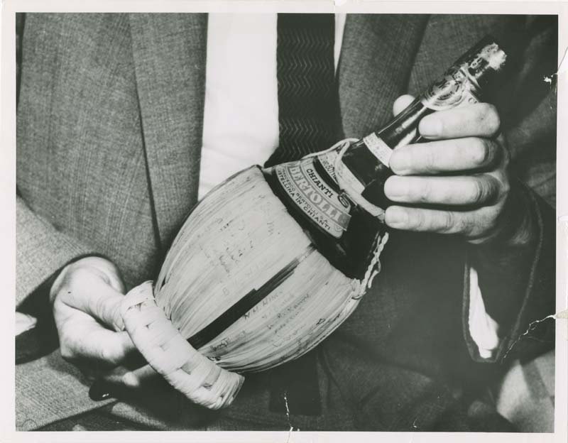 A black and white close-up photo of a bottle of Chianti champagne with a wooden sheath around the bottom of the bottle, held in a man's hands