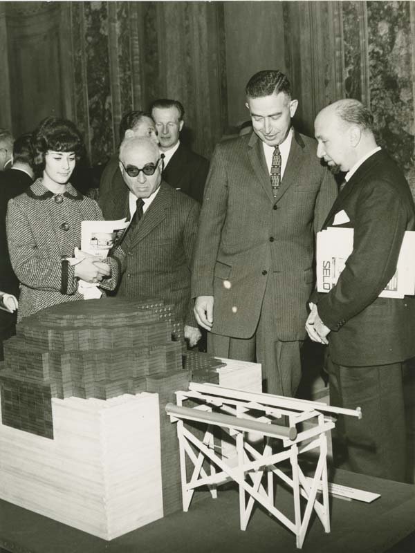 Three men in suits and a woman in a coat dress view a scale model of CP-1 at a twentieth anniversary celebration in Italy