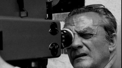 A black and white photograph of Luchino Visconti with his eye to the eyepiece of a camera.