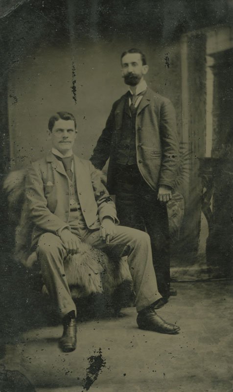 Black-and-white tintype formal photograph of Thomas E. Wilson seated next to a standing man. Thomas is a white man in his 20s or 30s, and is wearing a light-colored suit with a vest and wide tie. He is seated on a chair draped in an animal skin. To his left, another man is posed standing with his arm resting on the back of Thomas' chair. The standing man is a white man with a full dark beard, wearing a dark suit.