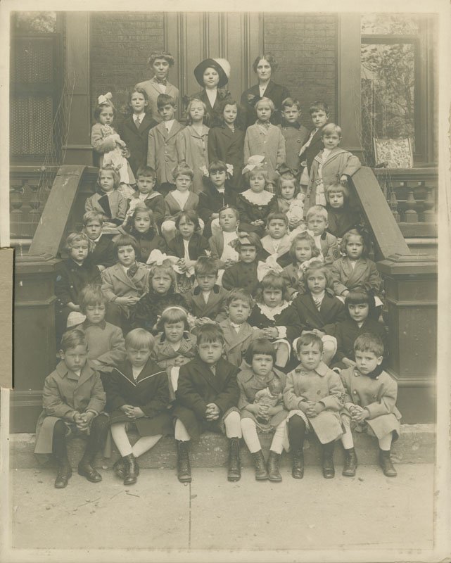 Black-and-white formal photograph of children seated and standing on front exterior steps of a brick building. Three adult women stand in the back row at the top of the stairs. The children are white boys and girls, approximately age four. Thomas E. Wilson stands in the back row, third from the right. He has a serious expression and is wearing a dark double-breasted coat with a darker collar.
