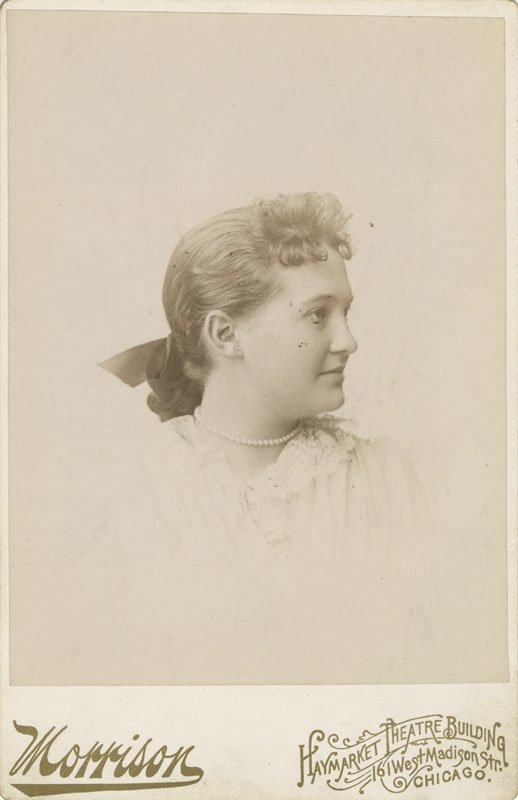 Black-and-white formal photographic portrait of Elizabeth Foss Wilson as a young girl. She is a young white woman shown from the shoulders up, wearing a light colored dress with short dark hair tied in a bow at the nape of her neck. She is shown in three-quarters profile.
