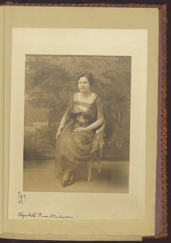 Black-and-white formal photographic portrait of Elizabeth Foss Wilson. She is shown seated in a chair in front of a backdrop that depicts a pastoral scene of trees and flowers. She is a middle-aged white woman wearing a long chiffon dress. Her dark hair is curled and pinned up. Her legs are crossed at the ankles and she is resting her left arm in her lap. Her right arm drapes over the arm of the chair.