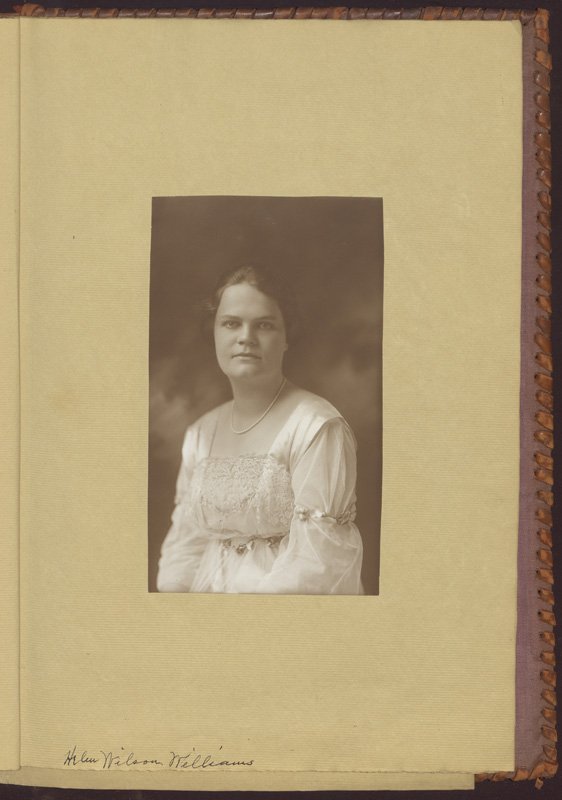 Black-and-white formal photographic portrait of Helen Wilson Williams pasted into a photograph album. Helen is a young white woman. She is wearing her dark hair pinned up, a light-colored dress, and a pearl necklace.