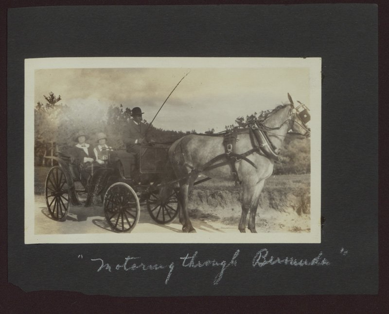 Black-and-white photograph of two white women (one of whom is Helen Wilson Williams) riding in a horse-drawn carriage driven by a Black man. Sandy ground and trees can be seen behind them. The photograph is attached to a black photo album page, and someone has written in white lettering "Motoring through Bermuda" below the photograph.