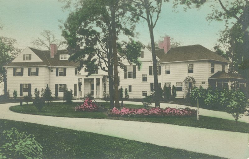 Color postcard of front of Edellyn Farm house. The house is a two-story, colonial mansion with white siding, black shutters, and a portico. There is a circle driveway in front of the house with trees and pink flowering bushes.