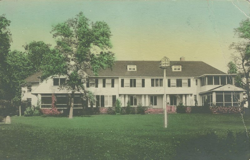 Color postcard of back of Edellyn Farm house. The house is a two-story, colonial mansion with white siding and black shutters. There is a large, grassy lawn behind the house, several trees, and a bird feeder on a tall pole.