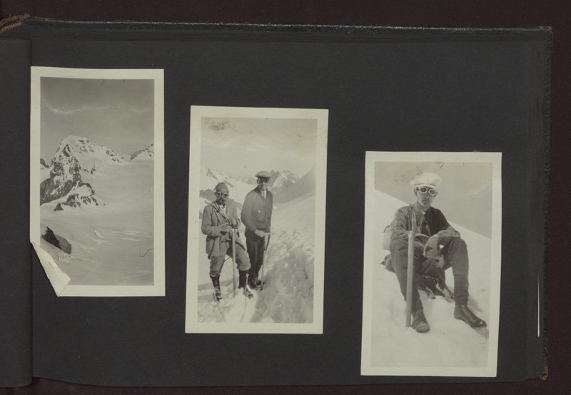 Three black-and-white photographs attached to a black photo album page. The first photograph is of a snowy mountain top. The second photograph is of Edward Foss Wilson and an unidentified man in climbing gear on a snowy mountain. The third photograph is of a man in climbing gear seated in the snow. They are all white men and are wearing goggles, caps, and boots, and are carrying ropes and pickaxes.
