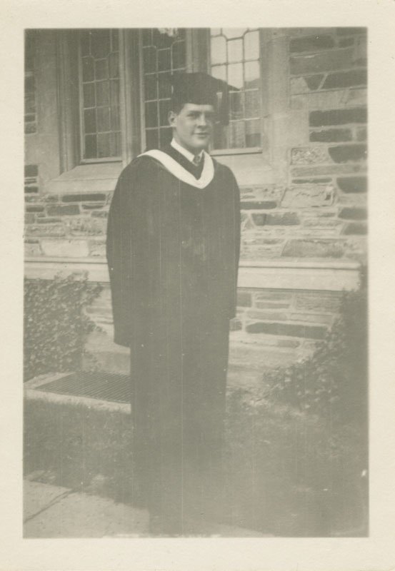 Black-and-white snapshot of Edward Foss Wilson wearing a graduation cap and gown. He is standing outdoors in front of a stone building.