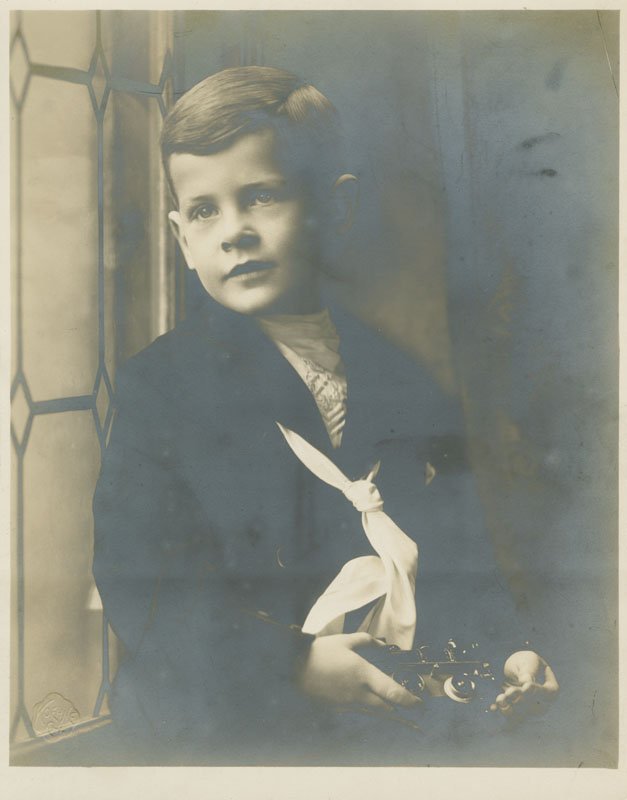Black-and-white formal photographic portrait of Edward Foss Wilson as a young child. Edward appears to be about four years old, has short dark hair, and is wearing a dark sailor suit. He is holding a toy train and is seated next to a window.
