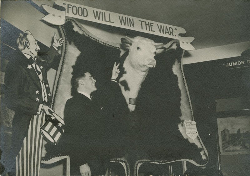 Black-and-white photograph of Edward Foss Wilson standing in front of a sign that reads "Food Will Win the War." He is raising his left hand in the "V for Victory" sign. Underneath the sign is a model of a cow. A cardboard cutout of Uncle Sam stands to Edward's right.