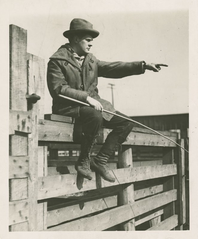 Black-and-white photograph of Edward Foss Wilson seated on a wooden fence rail in a stock yard. He is a young white man wearing a thick coat, laced boots, and a brimmed hat. He is holding a switch in his right hand and pointing straight ahead of him with his left hand.
