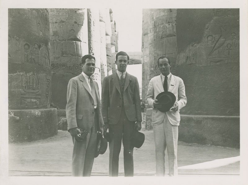 Black-and-white photograph of Edward Foss Wilson and two men in front of columns with hieroglyphics. All are white men wearing suits. They are each holding their fedora hat and looking into the camera.