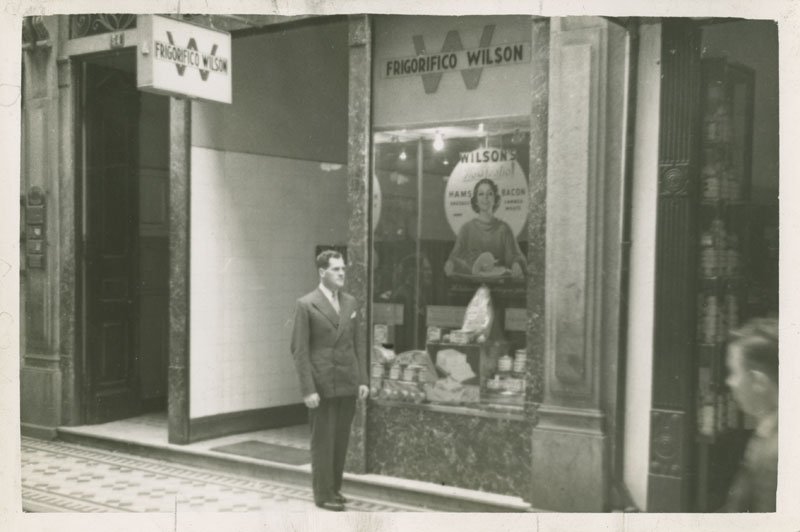 Black-and-white snapshot of Edward Foss Wilson standing outside in front of a building with a Wilson & Co. display in Brazil. He is a young white man with dark hair, and he is wearing a suit.