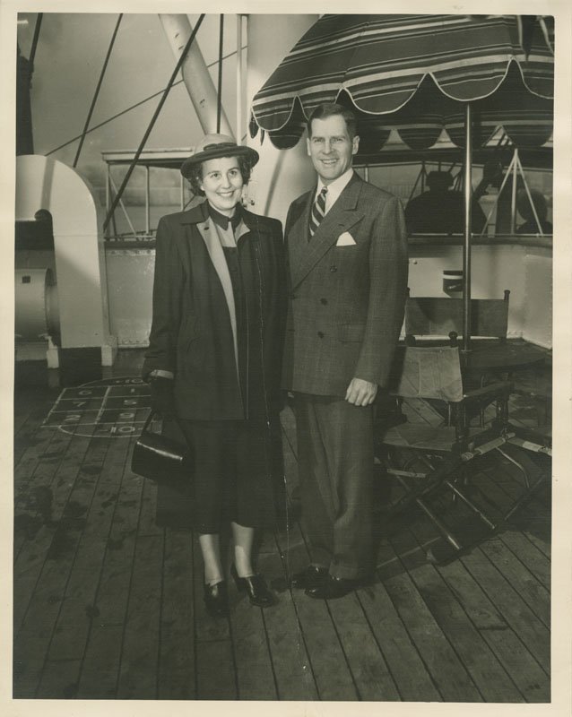 Black-and-white photograph of Pauline Wyman Wilson and Edward Foss Wilson standing on the deck of a cruise ship. Pauline is a young white woman wearing a dark skirt and coat with a hat and holding a purse. Edward Foss Wilson is a young white man and is standing next to Pauline wearing a dark suit. Behind them can be seen ship rigging, deck chairs and an umbrella, and a shuffleboard.
