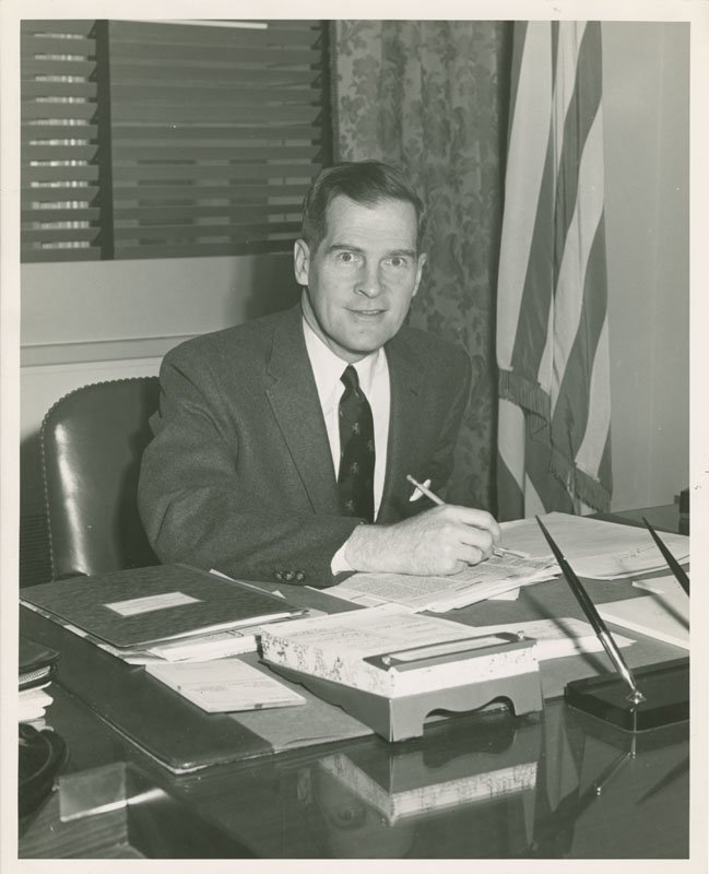 Black-and-white formal photograph of Edward Foss Wilson seated behind desk. He is a middle-aged white man wearing a dark suit and tie. He is holding a pen and writing on a document in front of him. He is looking into the camera. There is a window with blinds, and a draped American flag behind him.