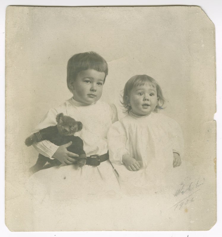 Black-and-white formal photographic portrait of Pauline Wyman Wilson as a baby and her older brother. Both are seated and shown from the waist up. Donald is a white boy with short dark hair. He is wearing a white shirt with a dark belt and is holding a teddy bear. Pauline is a white girl with short, dark, curly hair. She is wearing a white dress.