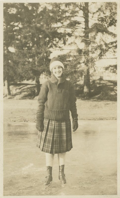 Black-and-white snapshot of Pauline Wyman Wilson ice skating outdoors. She is a young white woman and is wearing a plaid skirt, a knit sweater, and a knit hat. She is smiling facing the camera, and there are evergreen trees behind her.