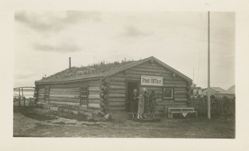 Black-and-white photograph of a log cabin structure with a "Post Office" sign above the front doorway. Three women in long coats and cloche hats stand at the front door of the building. Vegetation can be seen growing on the roof of the building.