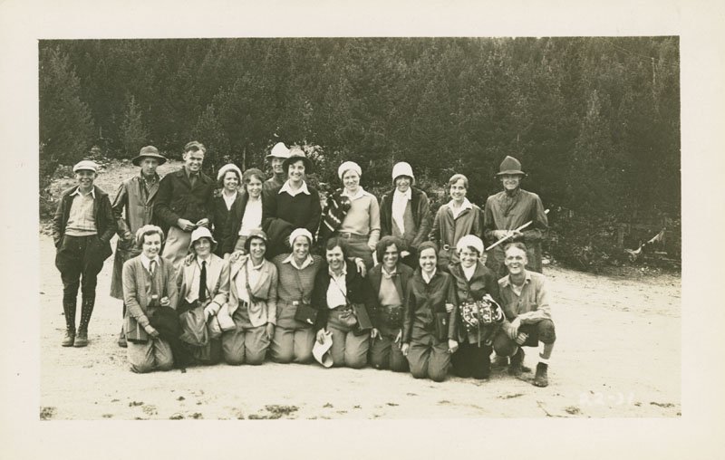 Black-and-white snapshot of a group of twenty young, white men and women. Most are wearing coats and hats, and are shown outdoors with pine trees behind them.