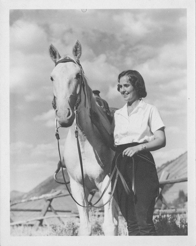 Black-and-white photograph of Pauline Wyman Wilson standing next to a horse. Pauline is a young white woman with short, dark, curly hair. She is smiling and holding the reigns of a white horse. Pauline is wearing a white shirt and dark trousers. They are standing in a fenced pasture with a mountain in the background.