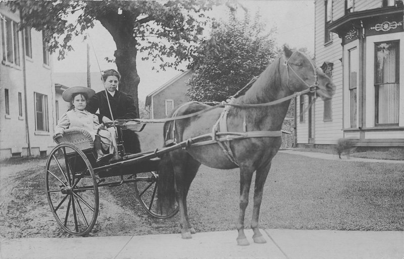 Black-and-white photographic postcard depicting Helen Wilson Williams as a young girl seated in a horse-drawn buggy. Helen's nurse stands next to her. The buggy is on a lawn and sidewalk between two white houses. Helen is a young white girl with long dark hair. She is wearing a light-colored coat and a light-colored brimmed hat.