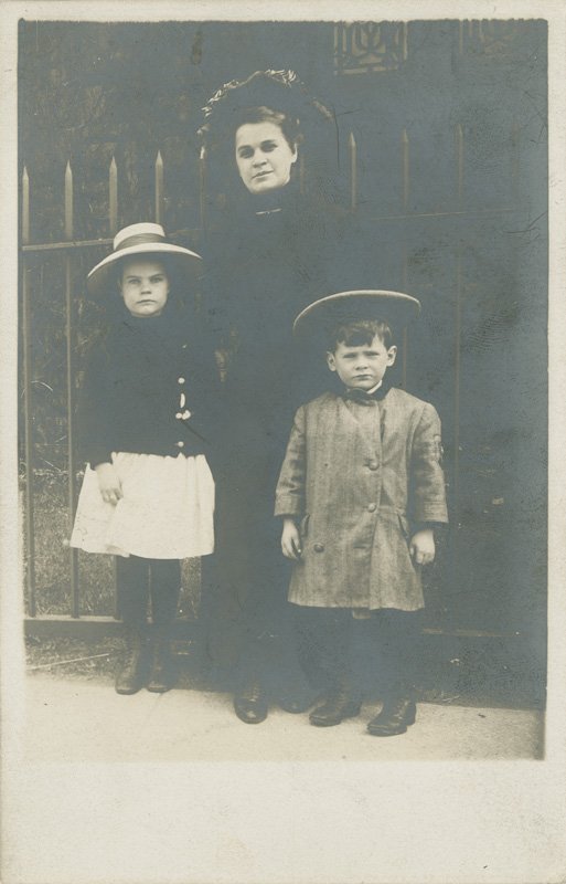 Black-and-white photographic postcard of Helen Wilson Williams as a young girl standing with her nurse Millie and younger brother, Edward. Helen is wearing a white skirt, white brimmed hat, dark sweater and stockings, and dark leather boots. She is white and has long dark hair.