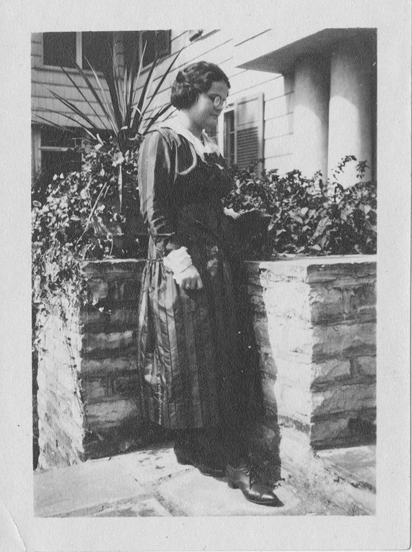 Black-and-white snapshot of Helen Wilson Williams as young woman standing outdoors on stone steps near planters. She is wearing a long dark dress and glasses. She is a young white woman with dark hair that is pinned up. She is turned sideways to the camera and is looking down.